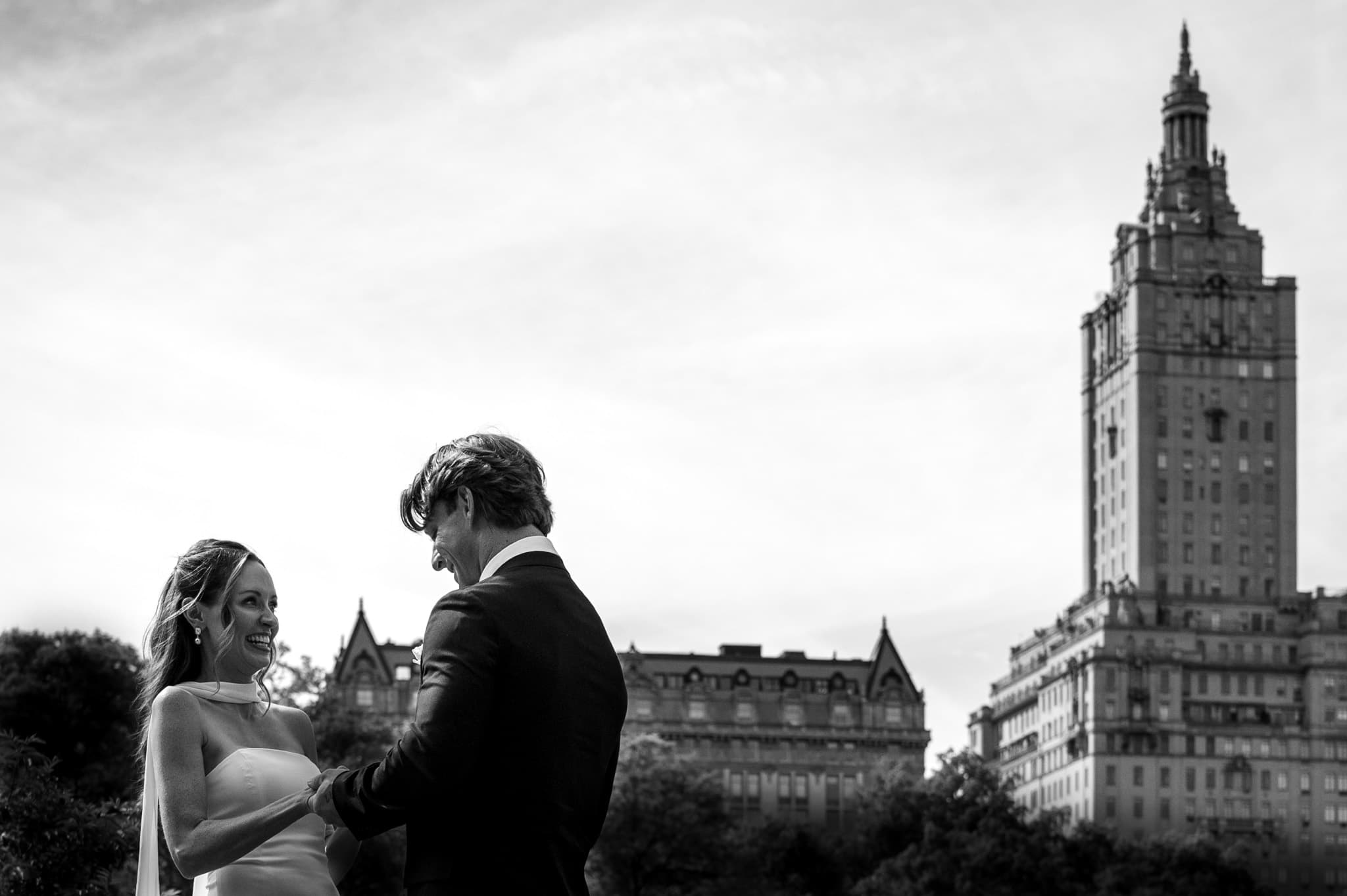 A couple in formal dress stand together at the edge of Central Park, the twin-towered San Remo and Upper West Side skyline rising behind the treeline.
