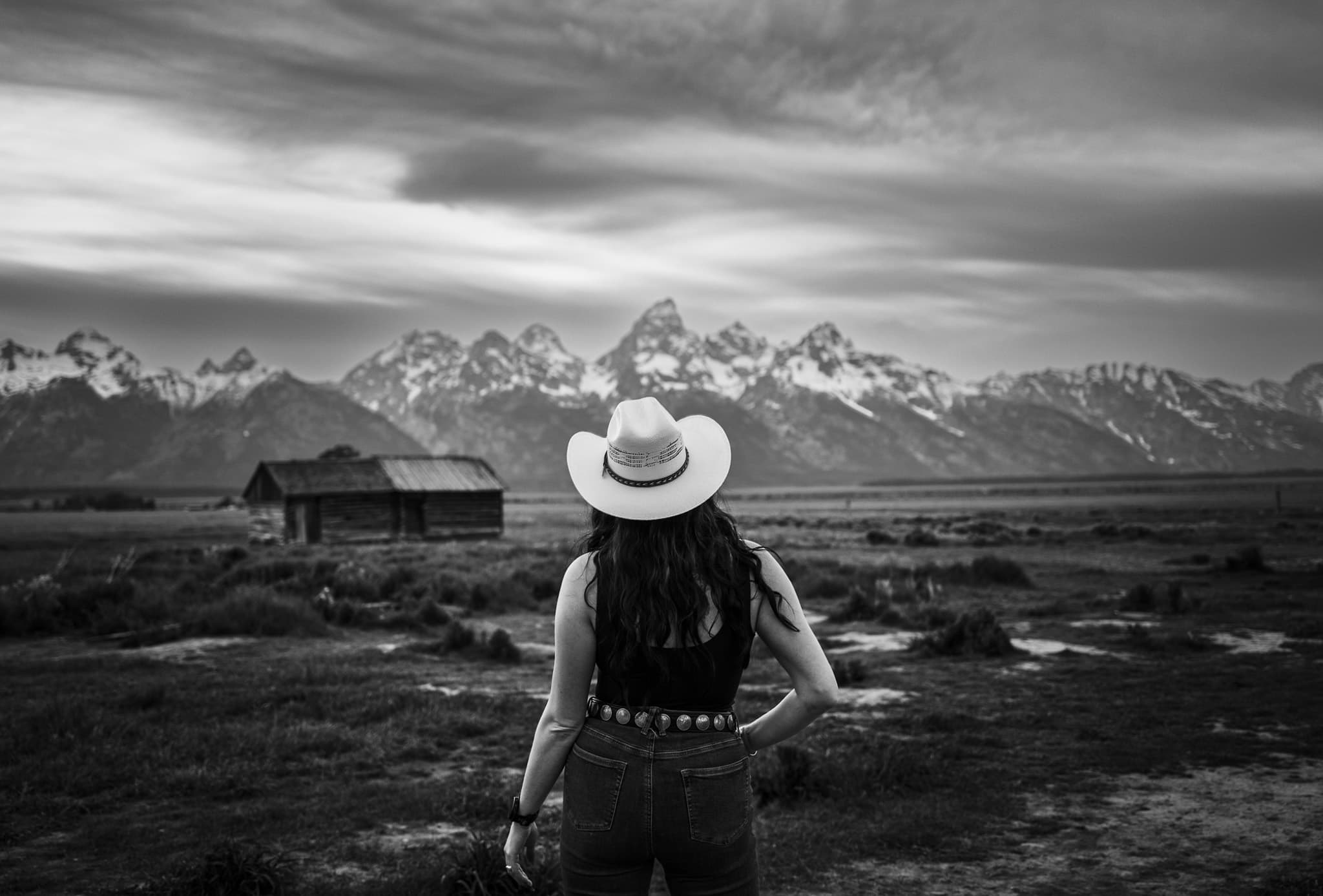 A figure in a wide-brimmed hat stands with their back to the camera before a snow-capped mountain range and a weathered timber cabin on an open plain.