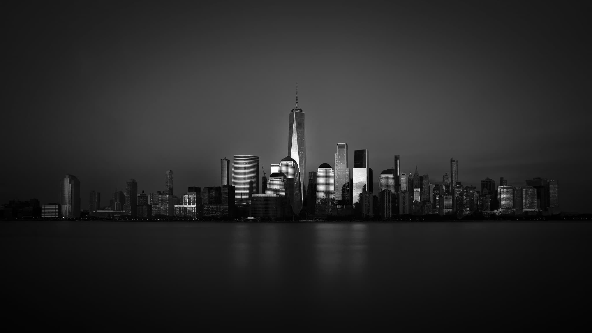 The Lower Manhattan skyline at dusk across calm water, One World Trade Center rising luminous at the center.