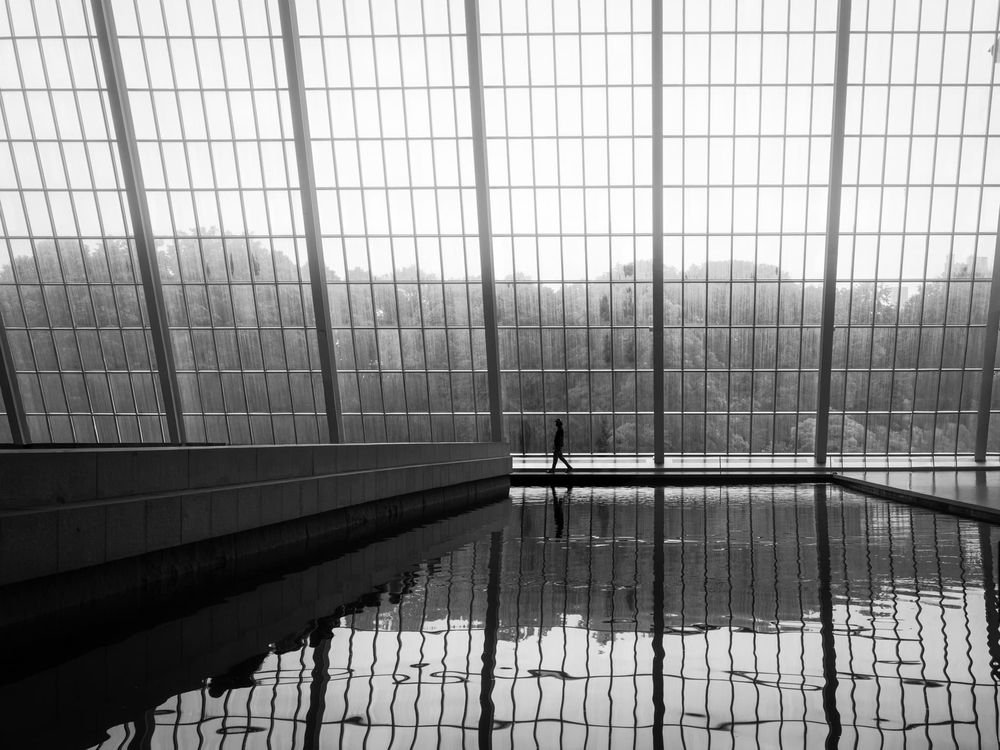 A lone figure crosses behind a still reflecting pool before the vast gridded glass wall of the Met's Sackler Wing, trees visible beyond.