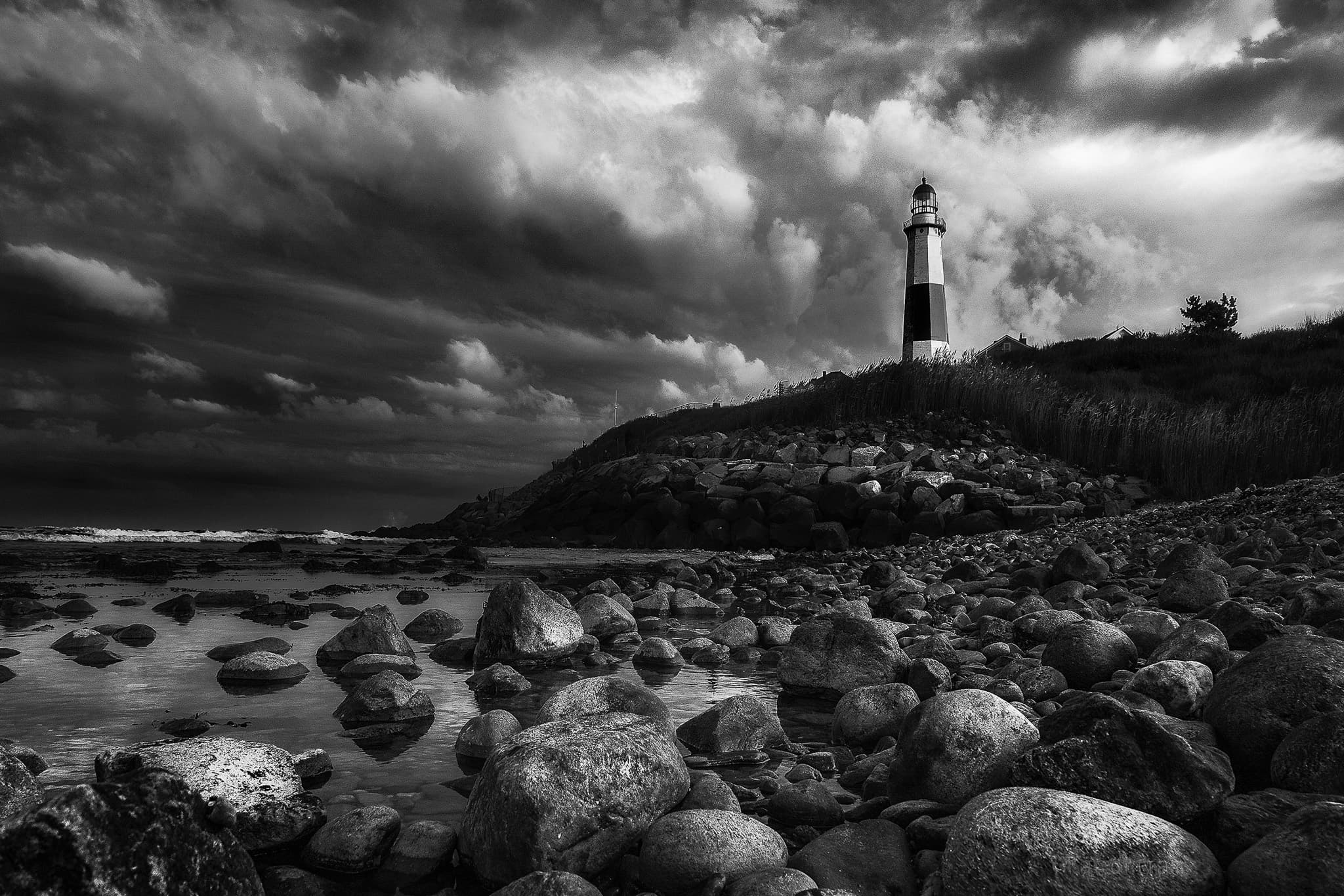 A black-and-white banded lighthouse above a boulder-strewn shore under a turbulent, dramatic sky.