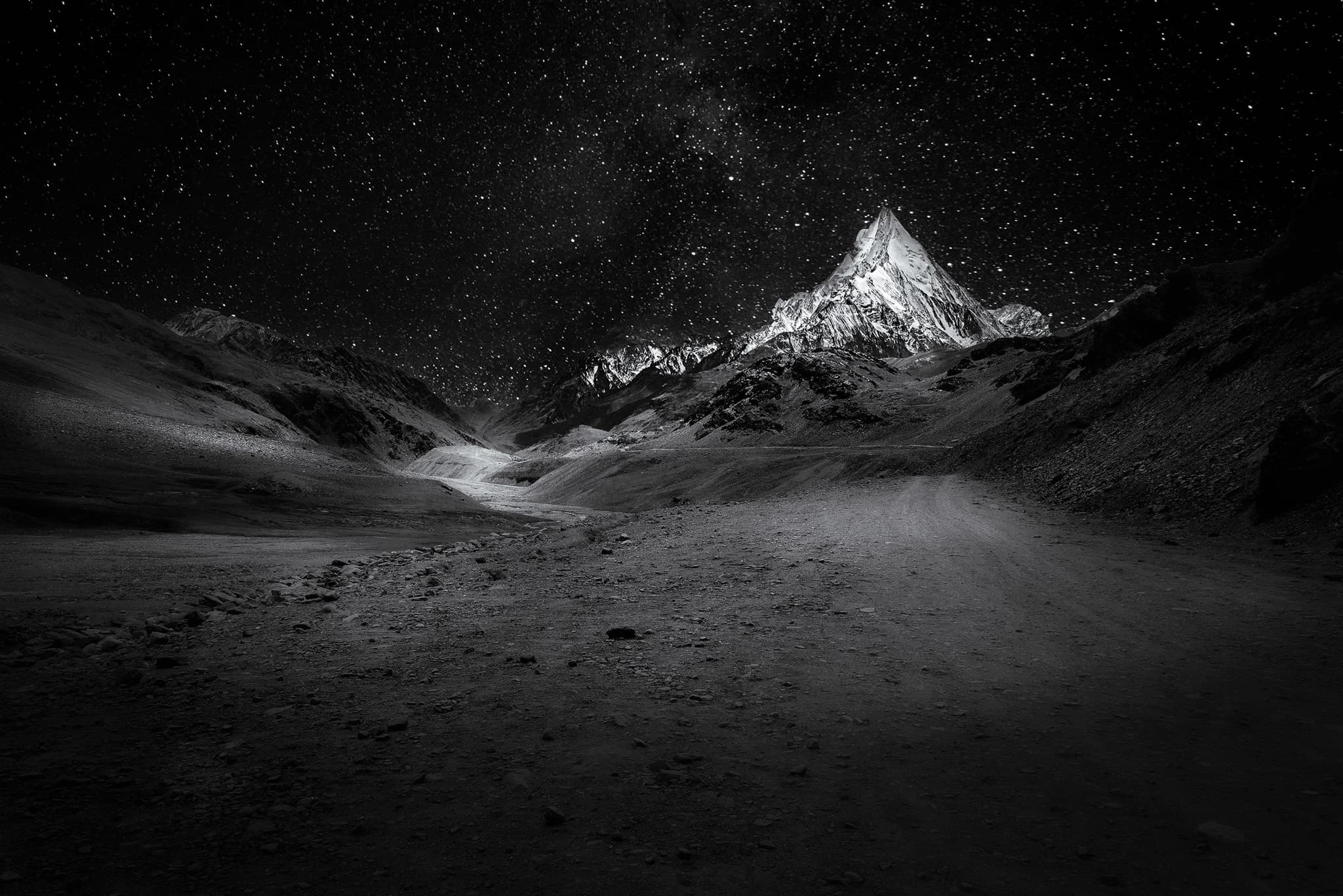 A snow-covered pyramidal peak catches faint light beneath a dense field of stars, above a dark gravel valley.