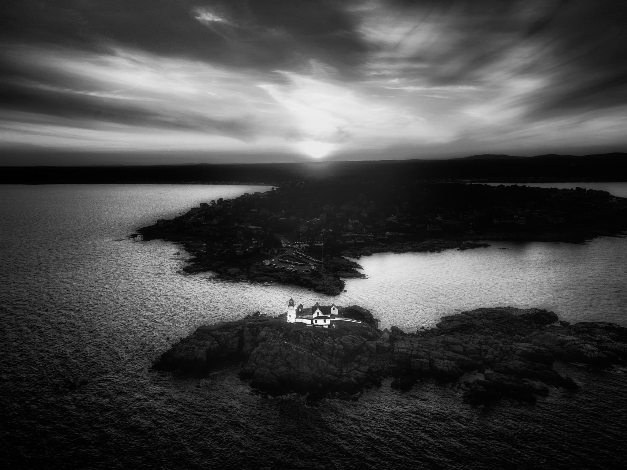 Aerial view of a clifftop lighthouse on a small rocky island at dusk, the sun low over a coastal village across the water.