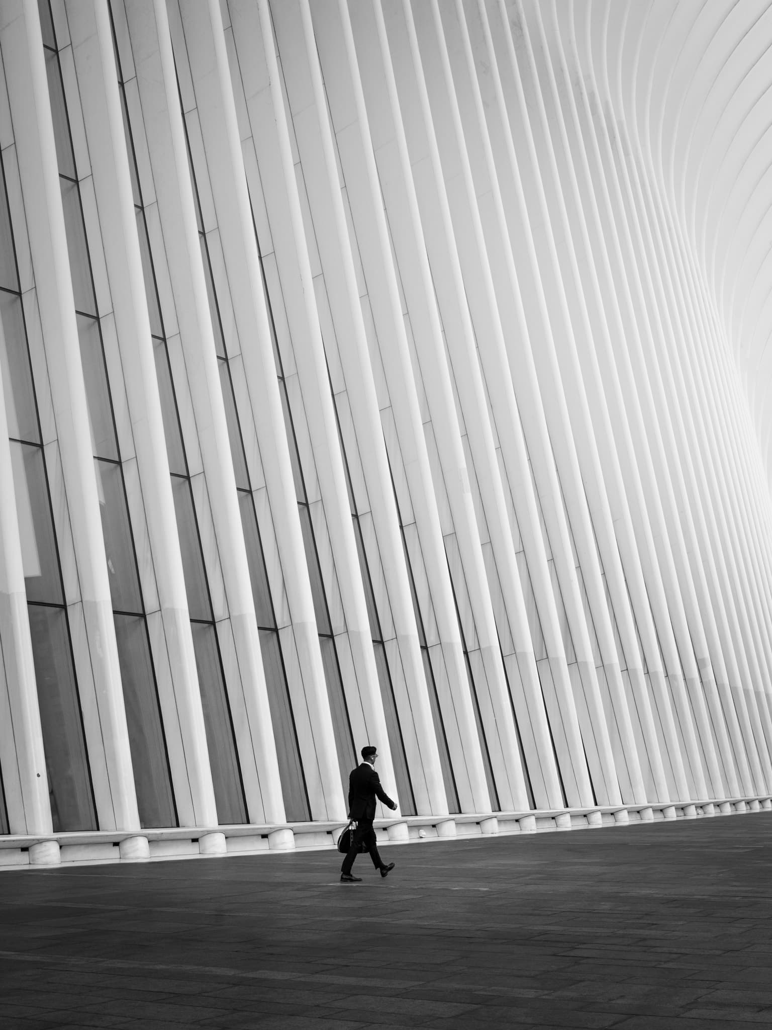 A lone figure in a dark suit walks past the soaring white ribbed wall of the Oculus transit hub, its louvers sweeping diagonally overhead.