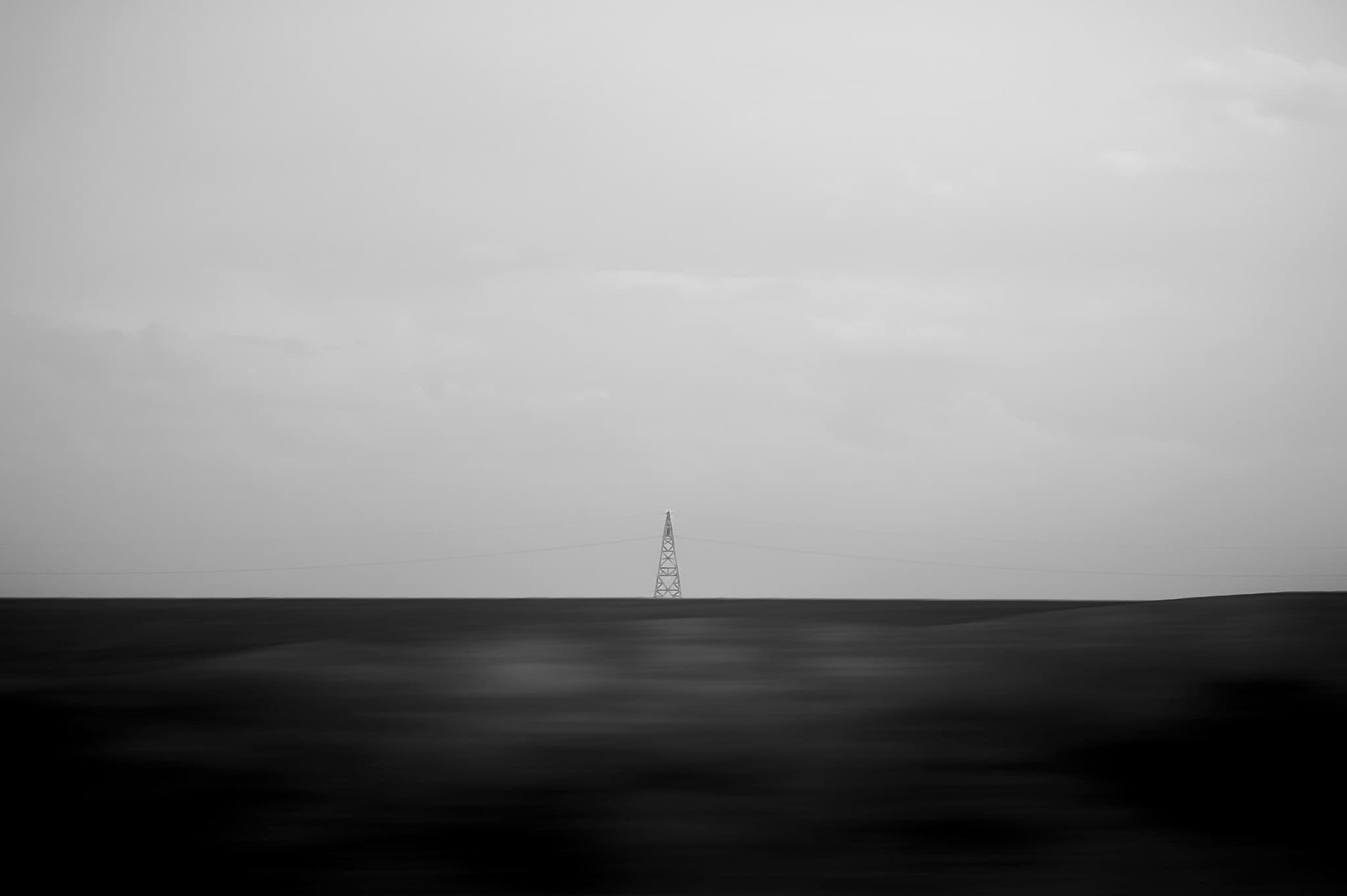 A single lattice transmission tower stands on the horizon above a dark, motion-blurred foreground under a pale sky — a near-abstract minimalist landscape.