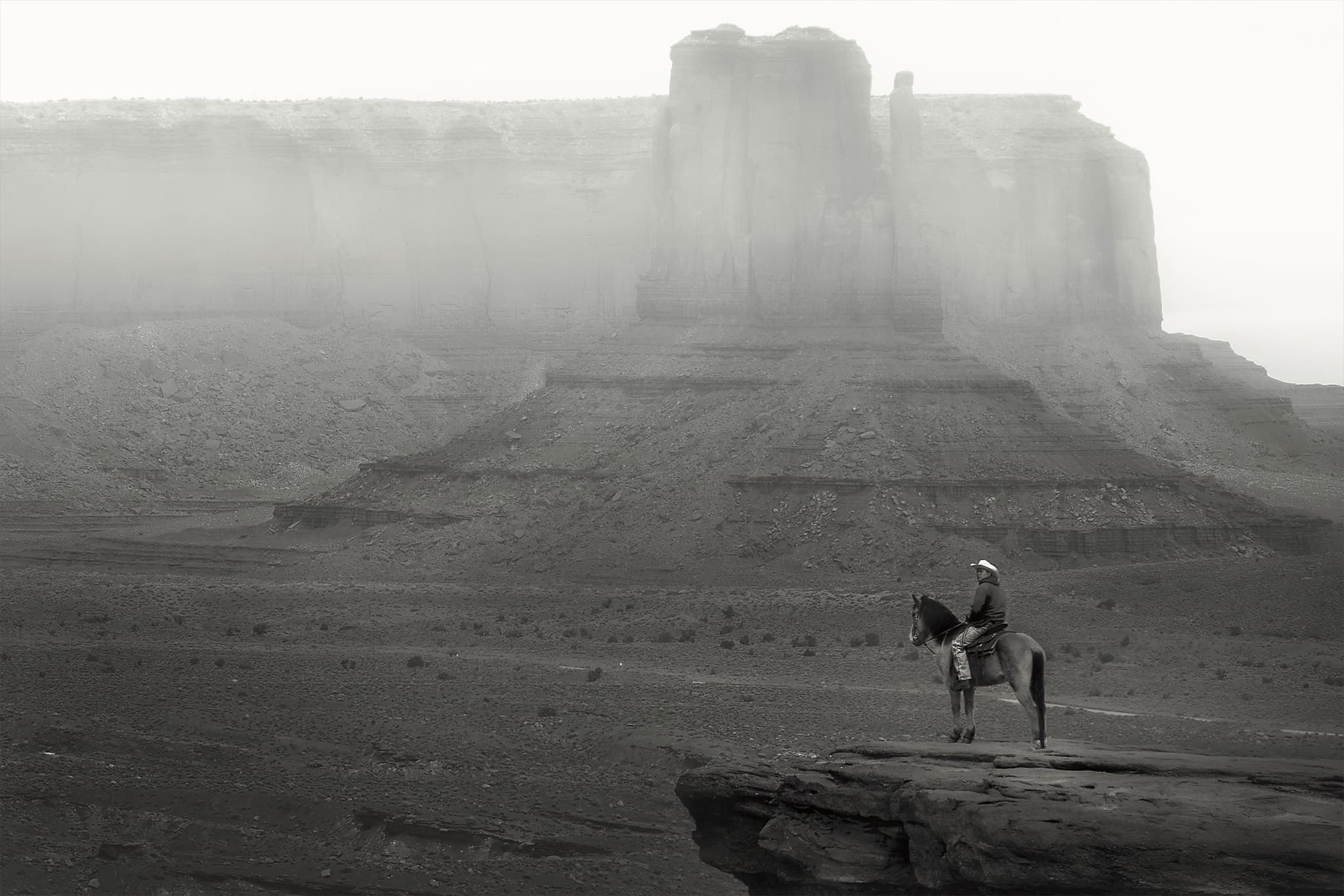 A lone horseman on a rock ledge faces a fog-shrouded sandstone mesa in the desert.