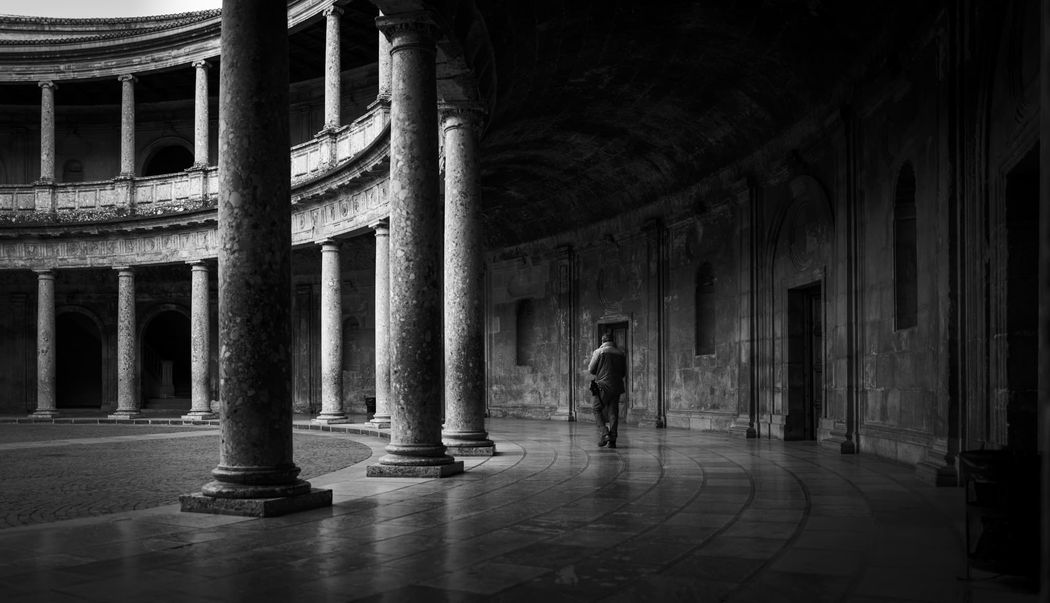 A lone figure crosses the circular two-tiered colonnaded courtyard of the Renaissance Palace of Charles V at the Alhambra, the curve of stone columns receding into shadow.