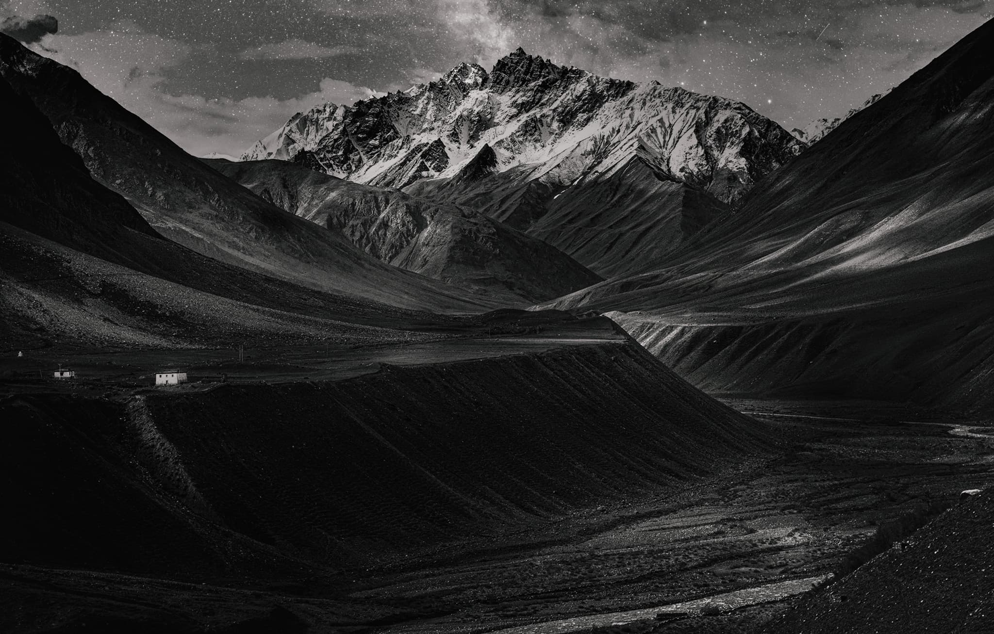 Snow-streaked mountains rise over a deep, dark high-altitude valley under a faintly starred sky, a few tiny buildings at the base for scale.