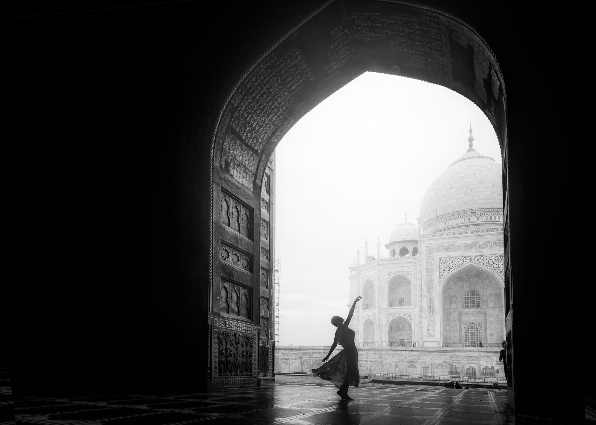 Framed by a dark carved archway, a dancer in motion is silhouetted against the white marble domes of the Taj Mahal at dawn.